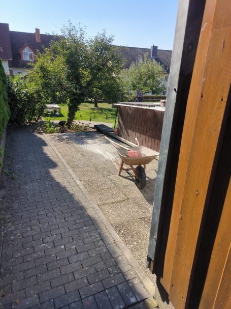 Gartenblick mit Schubkarre und Bäumen, sonniger Tag.
